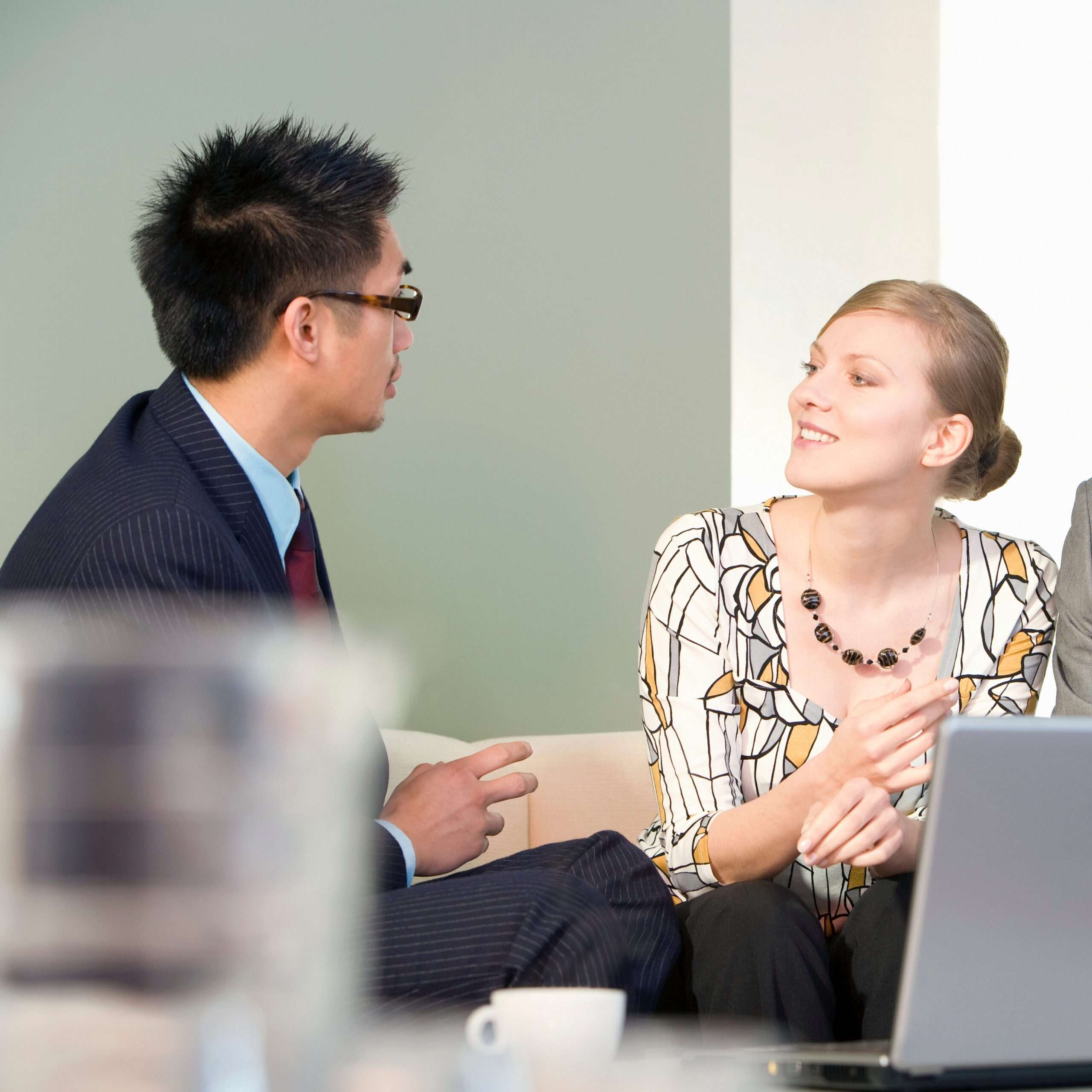 A man and a woman are engaged in a conversation in a professional setting. The man, wearing a suit and tie, faces the woman, who is smiling and holding a pen, dressed in a printed blouse. A laptop is open in front of them on a table.