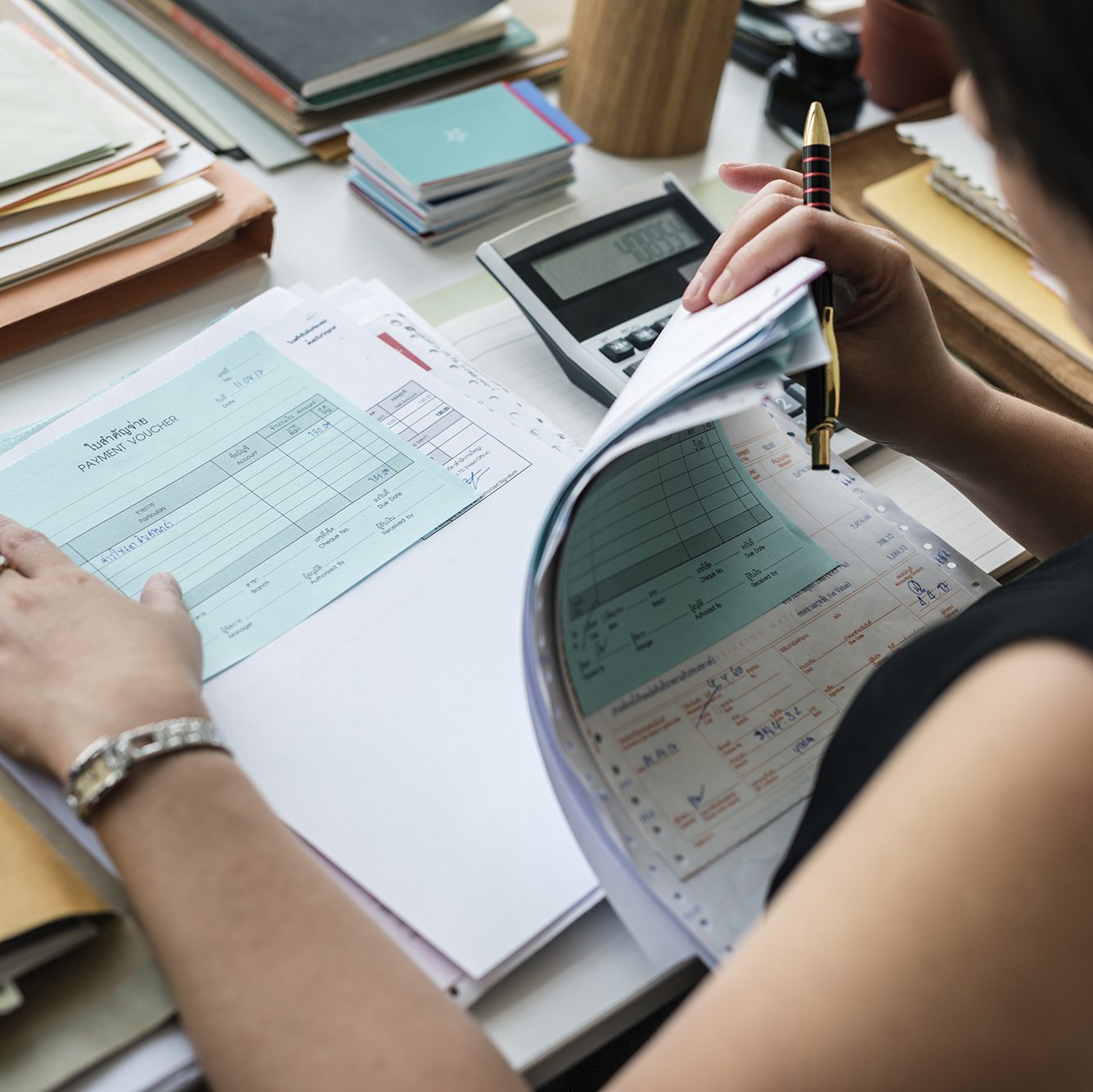 A person is holding a pen and examining paperwork at a desk. Various documents, including invoices and financial records, are spread out. A calculator, notebooks, and folders are also visible on the desk. The person appears to be focused on reviewing the documents.