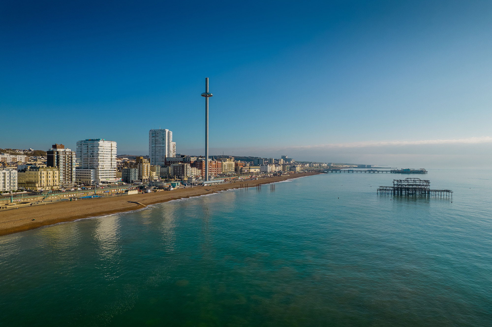 A coastal cityscape under a clear blue sky, featuring tall buildings, a prominent observation tower, a sandy beach, and a pier extending into calm, turquoise waters. The sun is casting a gentle glow over the scene.