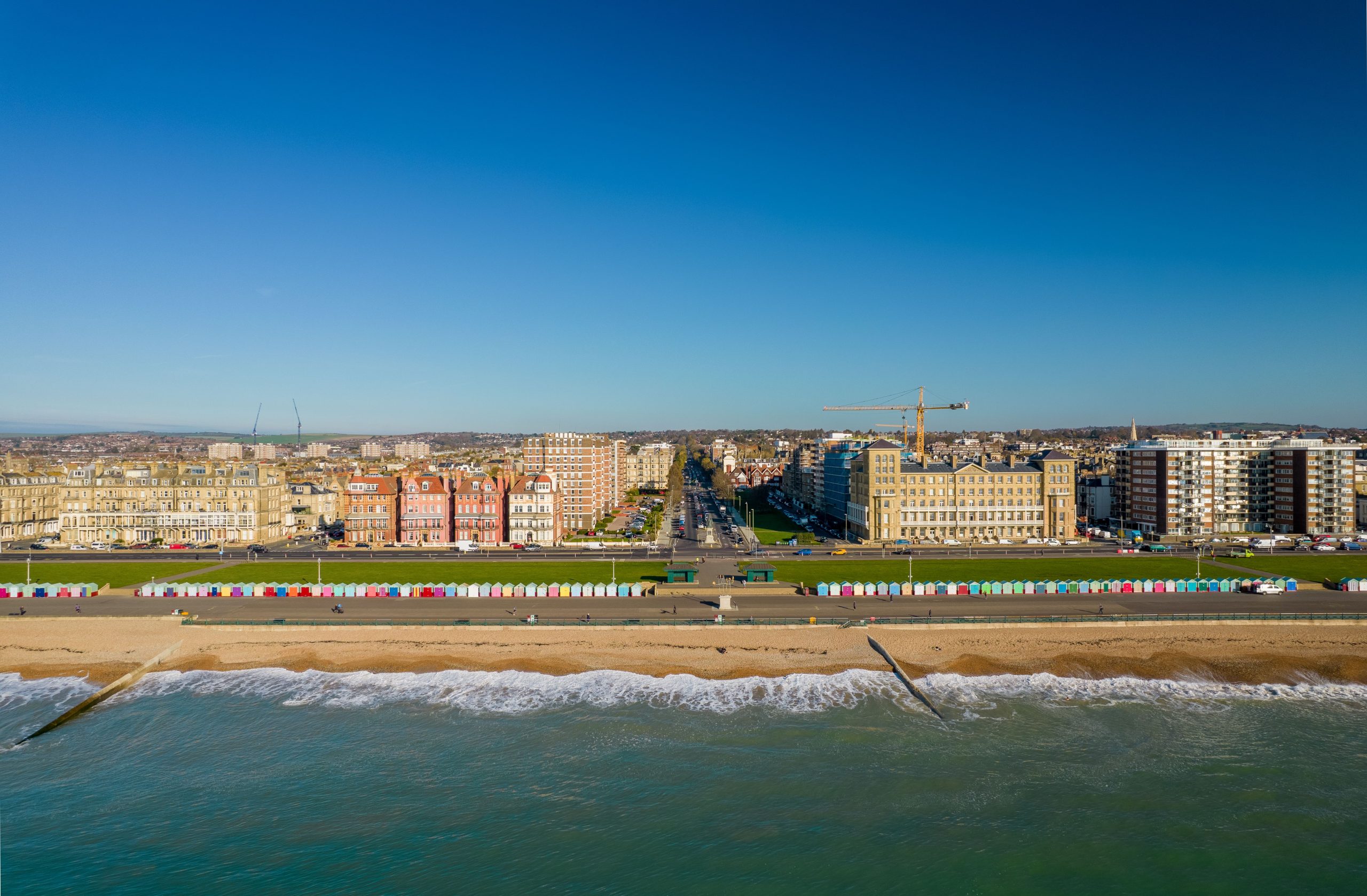 A coastal town with a sandy beach, colorful beach huts, and a promenade. Behind the promenade, there are rows of buildings including high-rise apartments and construction cranes. The sea is in the foreground and the sky is clear and blue.