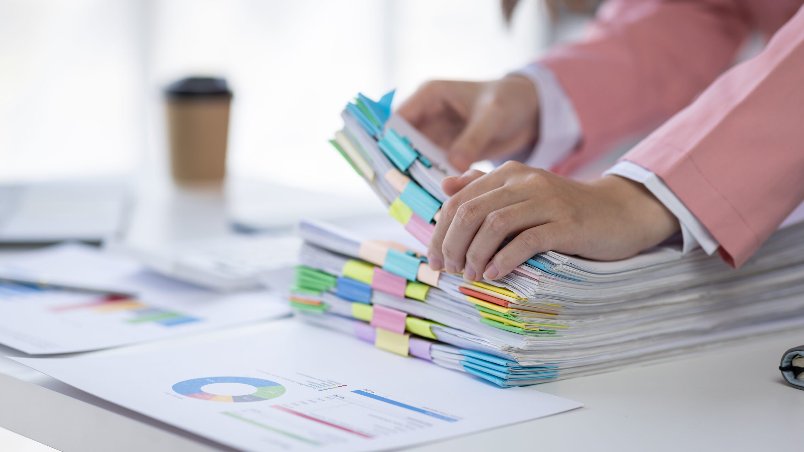 Two people in business attire sorting through a large stack of colorful, tabbed documents on a desk. Beside them are charts and a coffee cup, indicating they are organizing or reviewing business materials.