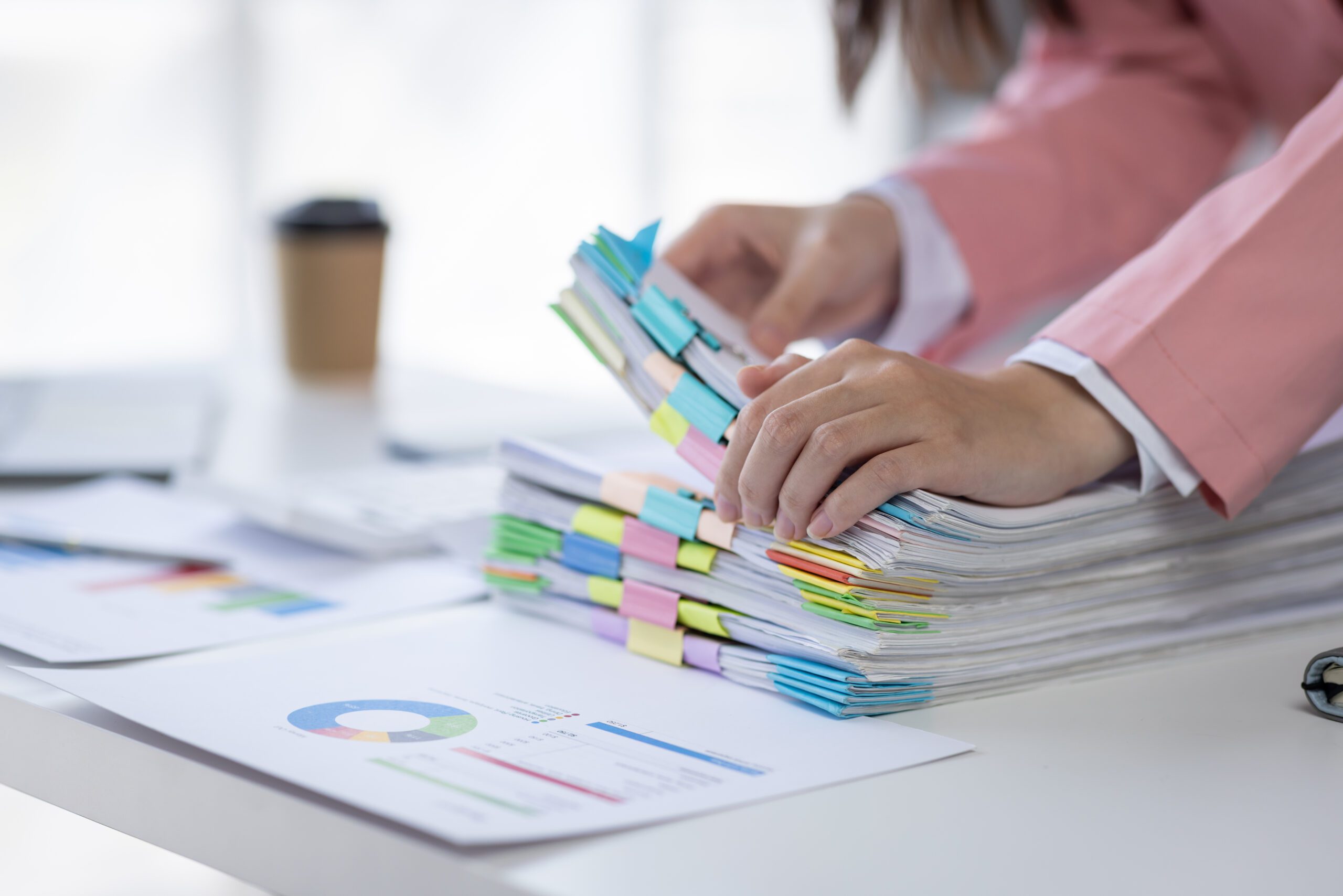 A person in a pink jacket is organizing a stack of papers held together with colorful paper clips on a bright, clean desk. Nearby, there is a report with a colorful pie chart, a keyboard, and a take-out coffee cup.