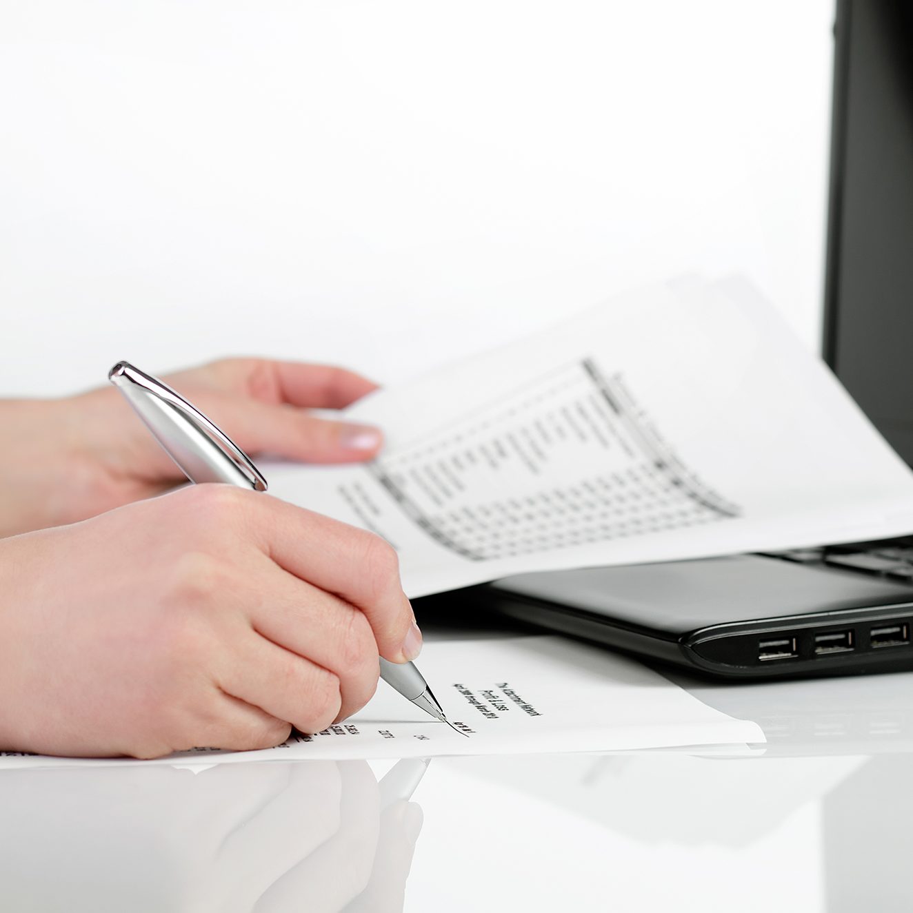 Person holding a pen and writing on a document while holding another sheet of paper, with a laptop nearby. The scene is set on a white desk and the focus is on the hands and papers, indicating a work or study environment.