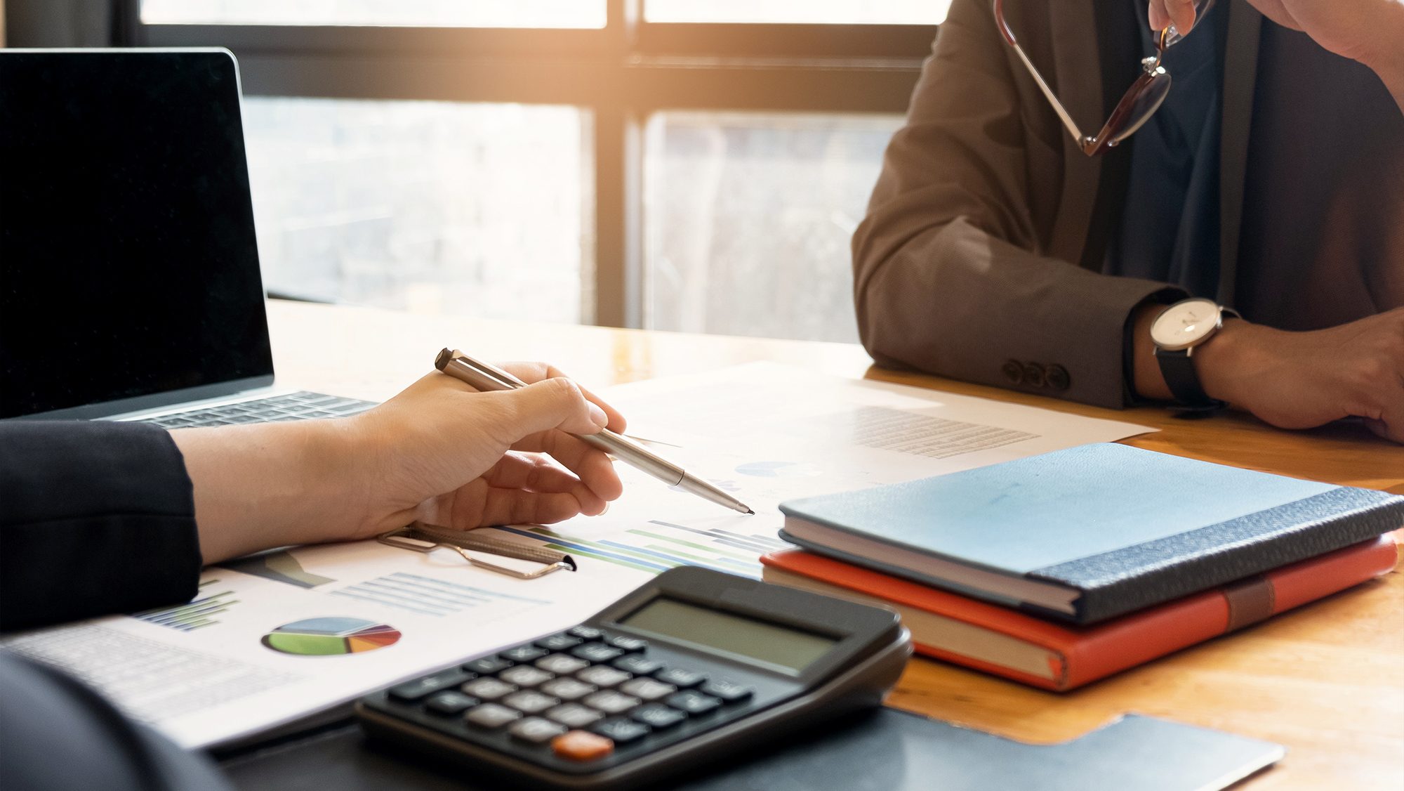 Two people sit at a wooden desk reviewing financial documents. One person points at graphs on a paper while holding a pen. A calculator, a laptop, and notebooks are also on the desk. The scene is lit by natural light from a window in the background.