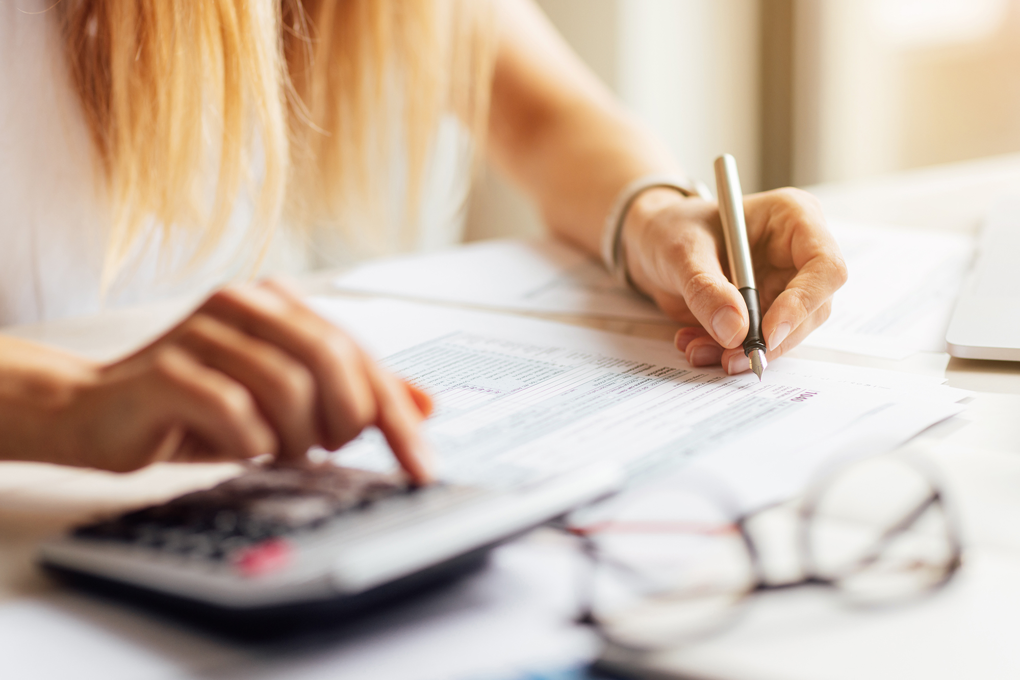 A person with long hair is sitting at a desk, holding a pen and writing on papers spread out in front of them. Their other hand is pressing buttons on a calculator. Eyeglasses are placed on the desk next to the papers.