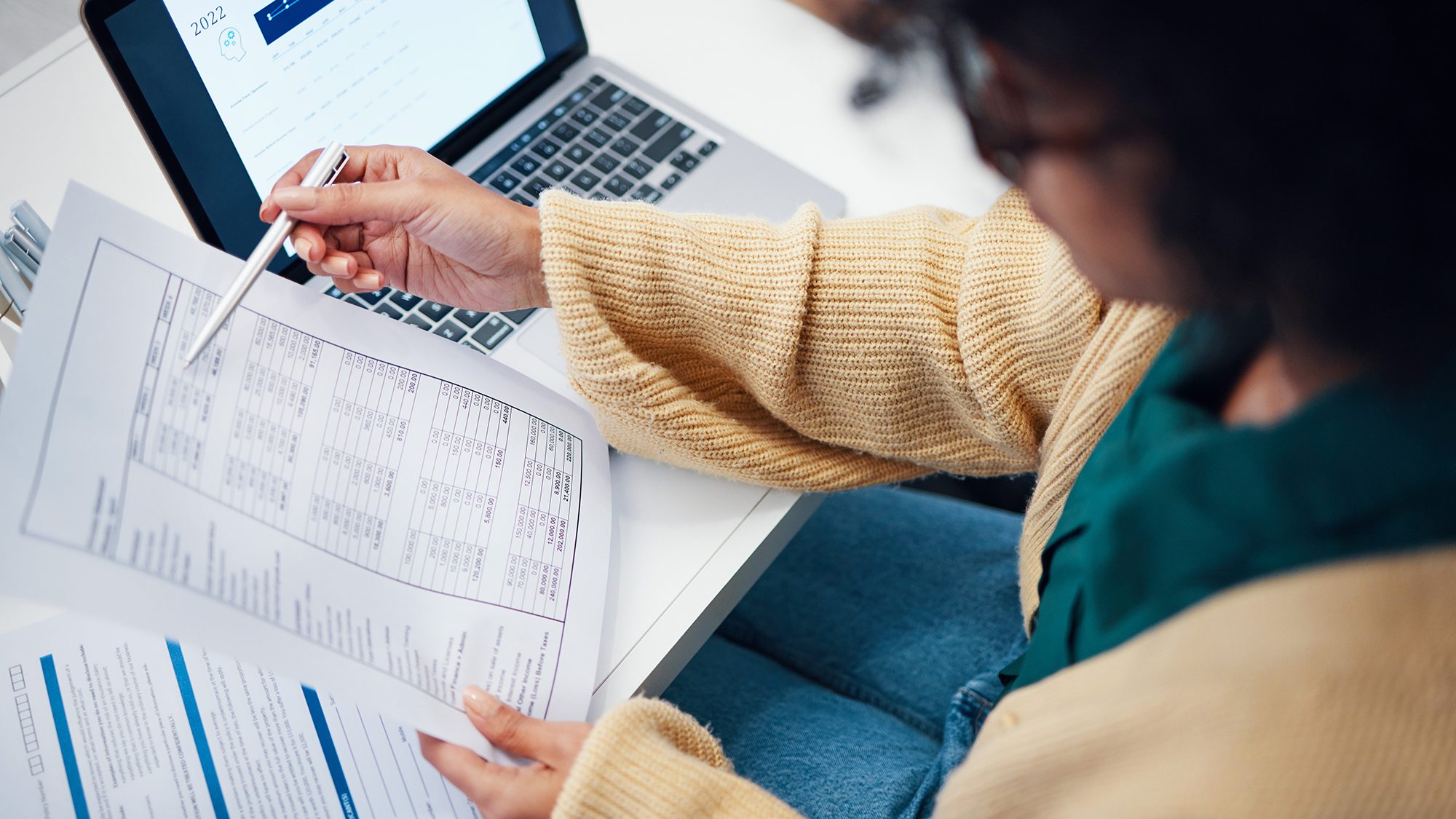 A person wearing a beige sweater and glasses is holding a pen and pointing at a printed document. They are seated at a desk with an open laptop displaying a webpage in the background. Several other documents are on the desk.