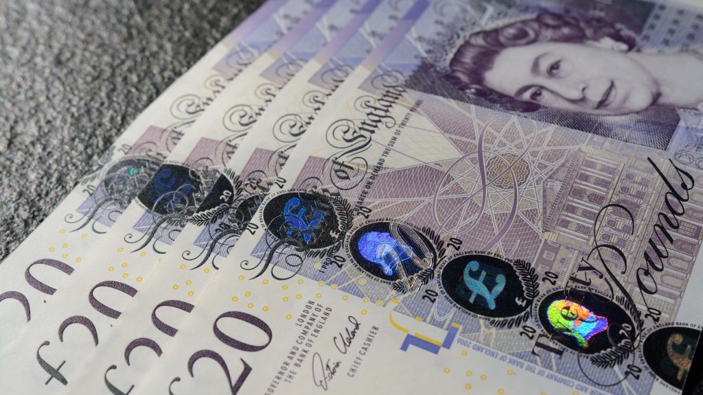 Close-up image of several British 20-pound banknotes fanned out on a textured surface. The notes feature a holographic stripe and a portrait of Queen Elizabeth II.