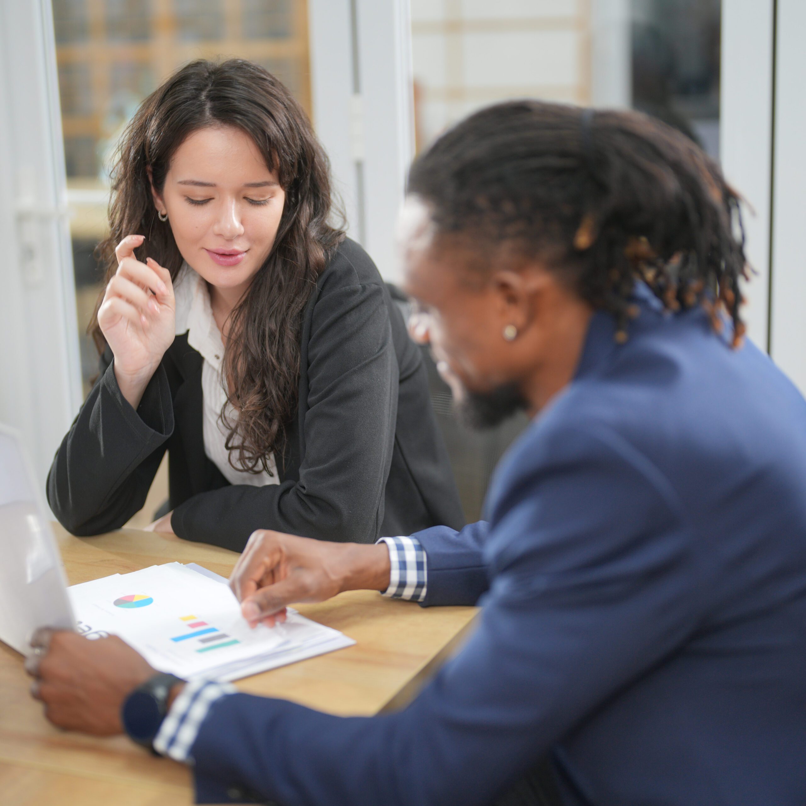 Two people, a woman and a man, are seated at a table in an office setting. They are engaged in a conversation while reviewing documents and charts. The woman is holding a pen, and the man is pointing at the papers. Both are dressed in business attire.