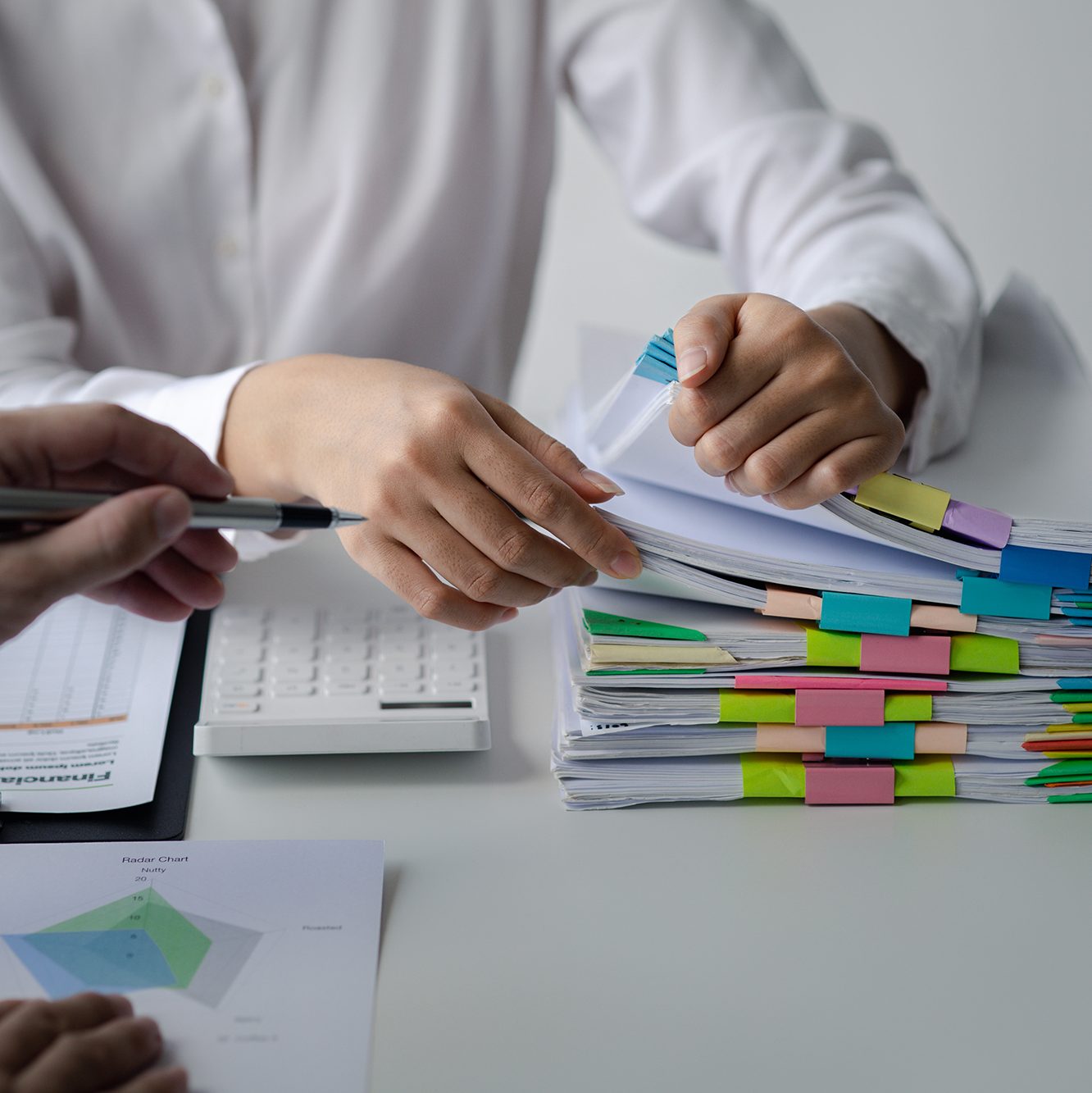 Two people are working together at a desk filled with a stack of documents organized with colorful paper clips. One person is pointing at a document with a pen, and the other is flipping through papers. A calculator and printed graphs are also visible on the desk.