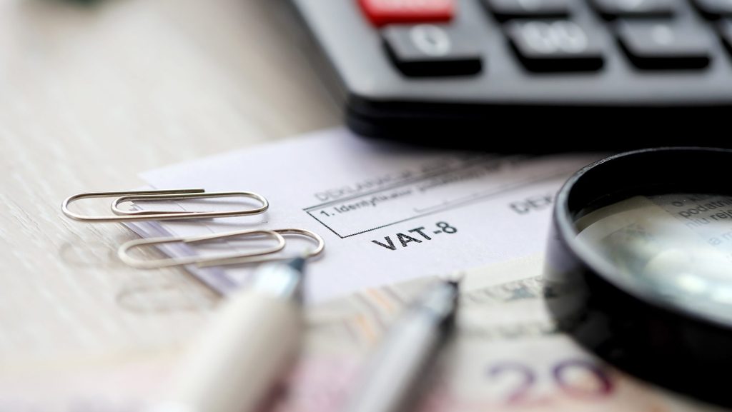 A close-up of a desk containing a financial document labeled "VAT-8" partially covered by two paper clips. A magnifying glass, pen, various currency notes, and a black calculator are also visible in the background.