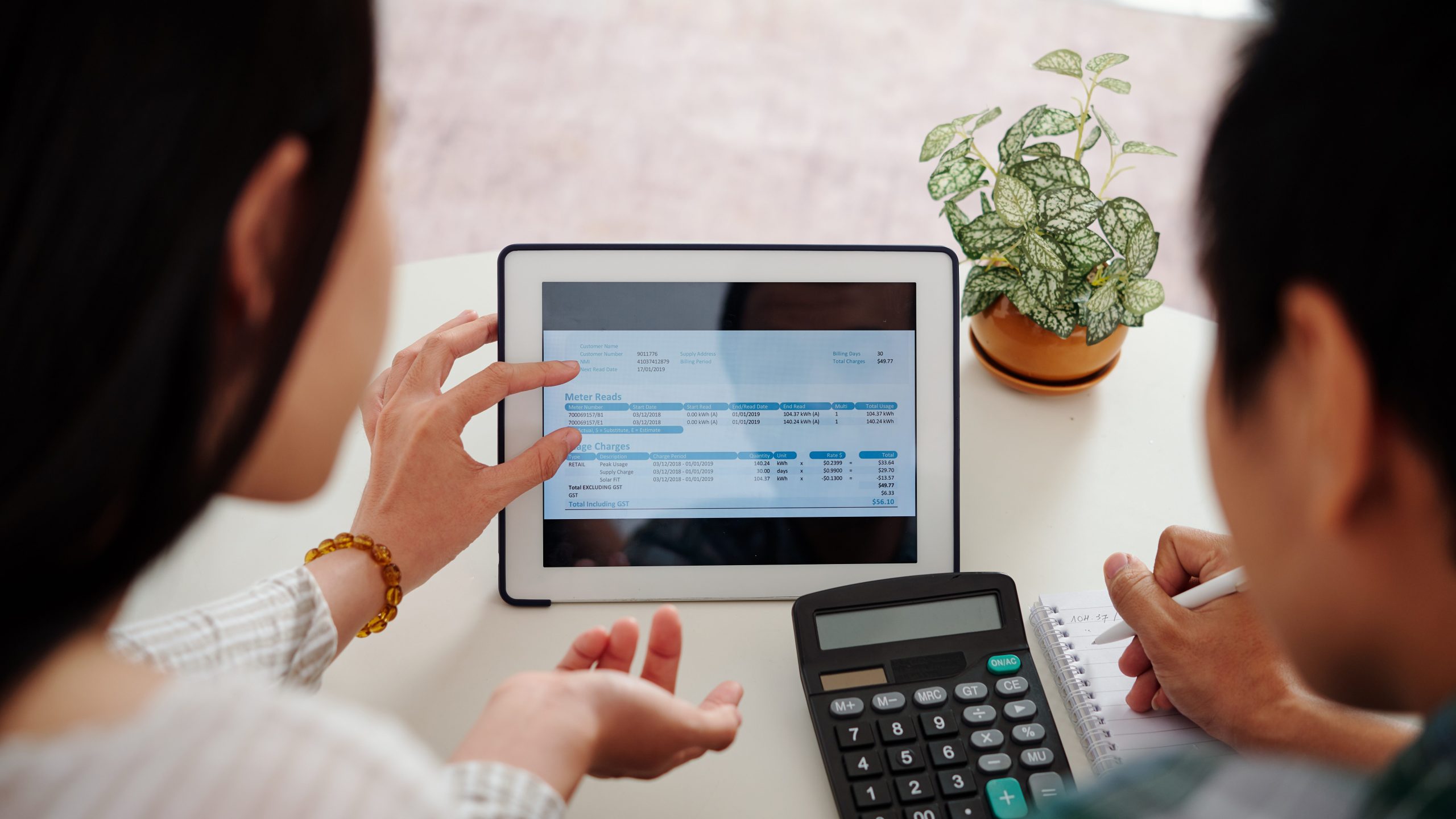 Two people are looking at a tablet displaying financial documents. One person is pointing at the screen, while the other is taking notes on paper. A calculator is placed on the table, and a small potted plant is visible in the background.