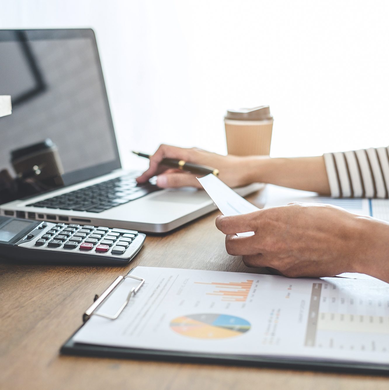 A person working at a desk with a laptop, holding a piece of paper in one hand, and typing with the other. A coffee cup, calculator, and documents with charts and graphs on a clipboard are also on the desk. The scene suggests a focus on financial work or analysis.