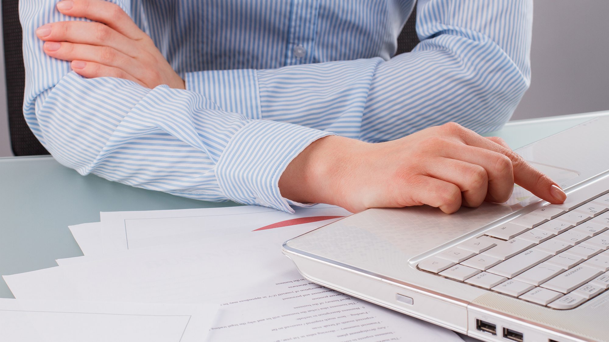 A person in a blue striped shirt is sitting at a desk with their arms crossed, using their right hand to type on a white laptop keyboard. Several sheets of paper are scattered on the desk.