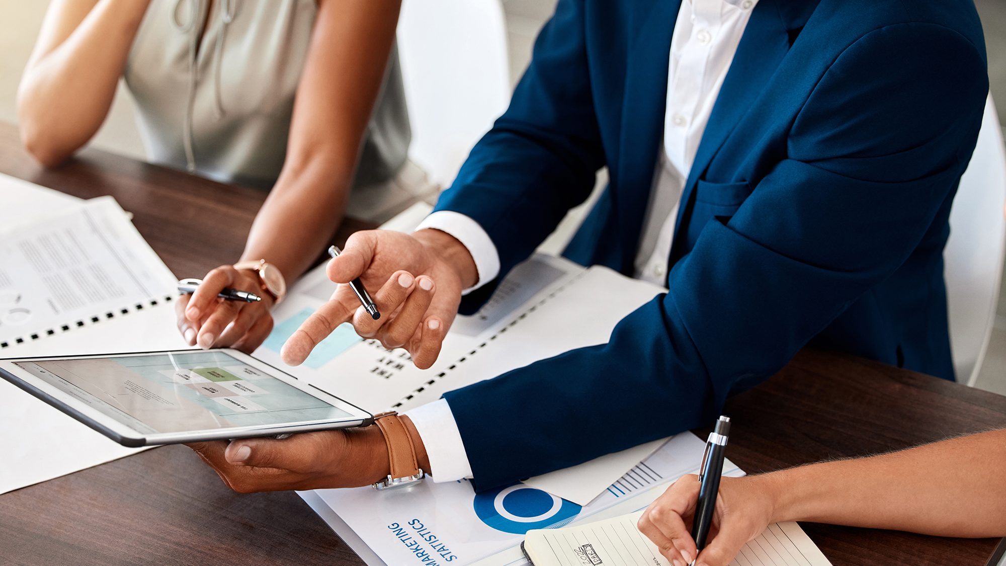 Three people are gathered around a wooden table, two of them pointing at a tablet screen with business documents and charts spread out. They are reviewing the content on the tablet, suggesting a discussion or meeting regarding the information displayed.