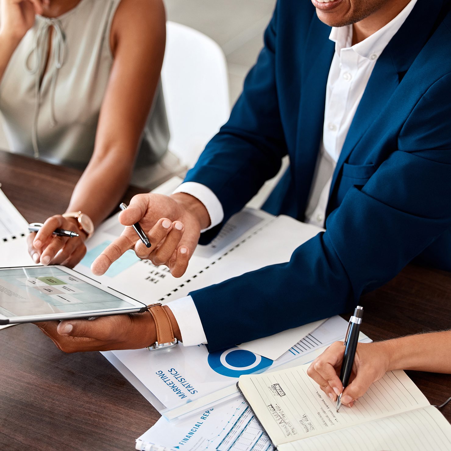 Close-up of three business professionals collaborating at a desk. One person in a blue suit points at a tablet, while another writes in a notebook and the third holds a document. Papers, charts, and notebooks are spread out on the table.