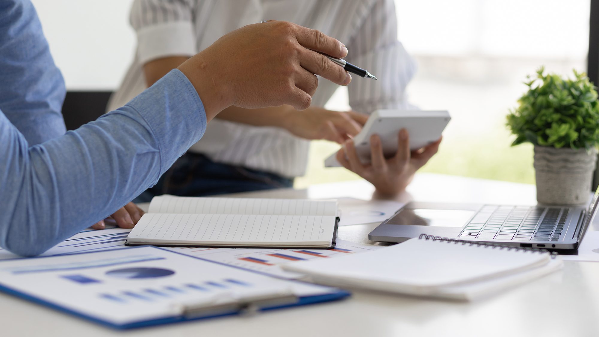 Two people are working at a desk with office supplies. One person is pointing at a notebook with a pen, while the other holds a tablet. There are documents, a laptop, a notebook, and a potted plant on the desk. Both are dressed in casual business attire.