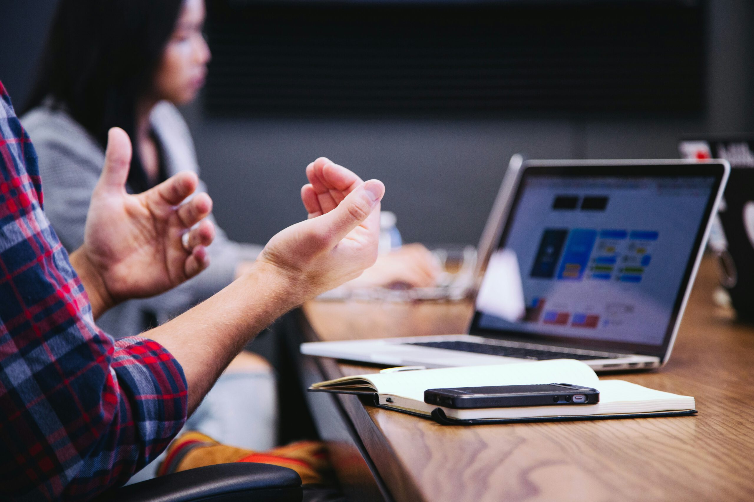 A person gesturing with their hands while discussing something. Beside them is a laptop displaying charts and a notebook with a pen and smartphone on a wooden table. Another person is sitting in the background, slightly out of focus.