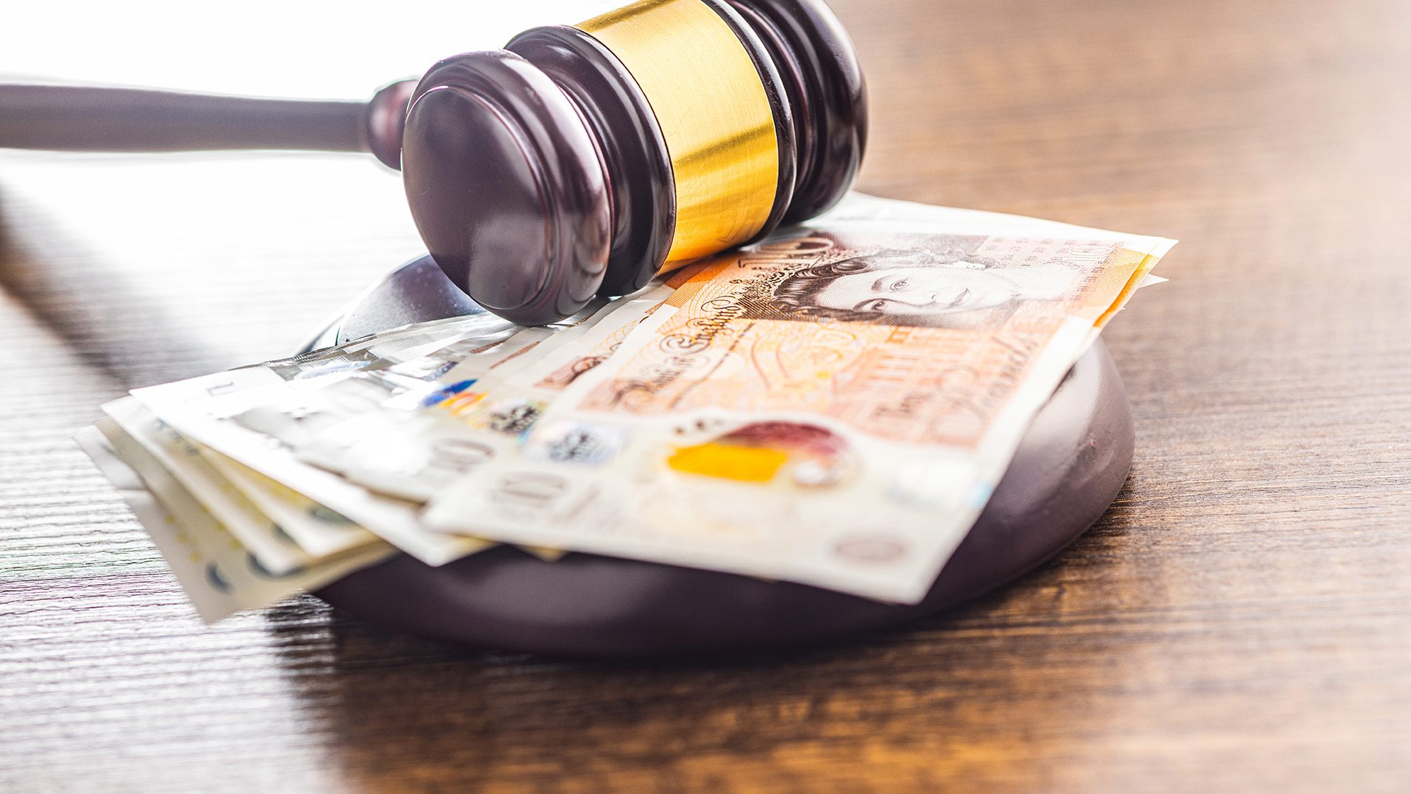 A wooden gavel rests on a stack of British paper currency, including five and ten-pound notes, on a polished wooden surface, symbolizing the intersection of law and finance.