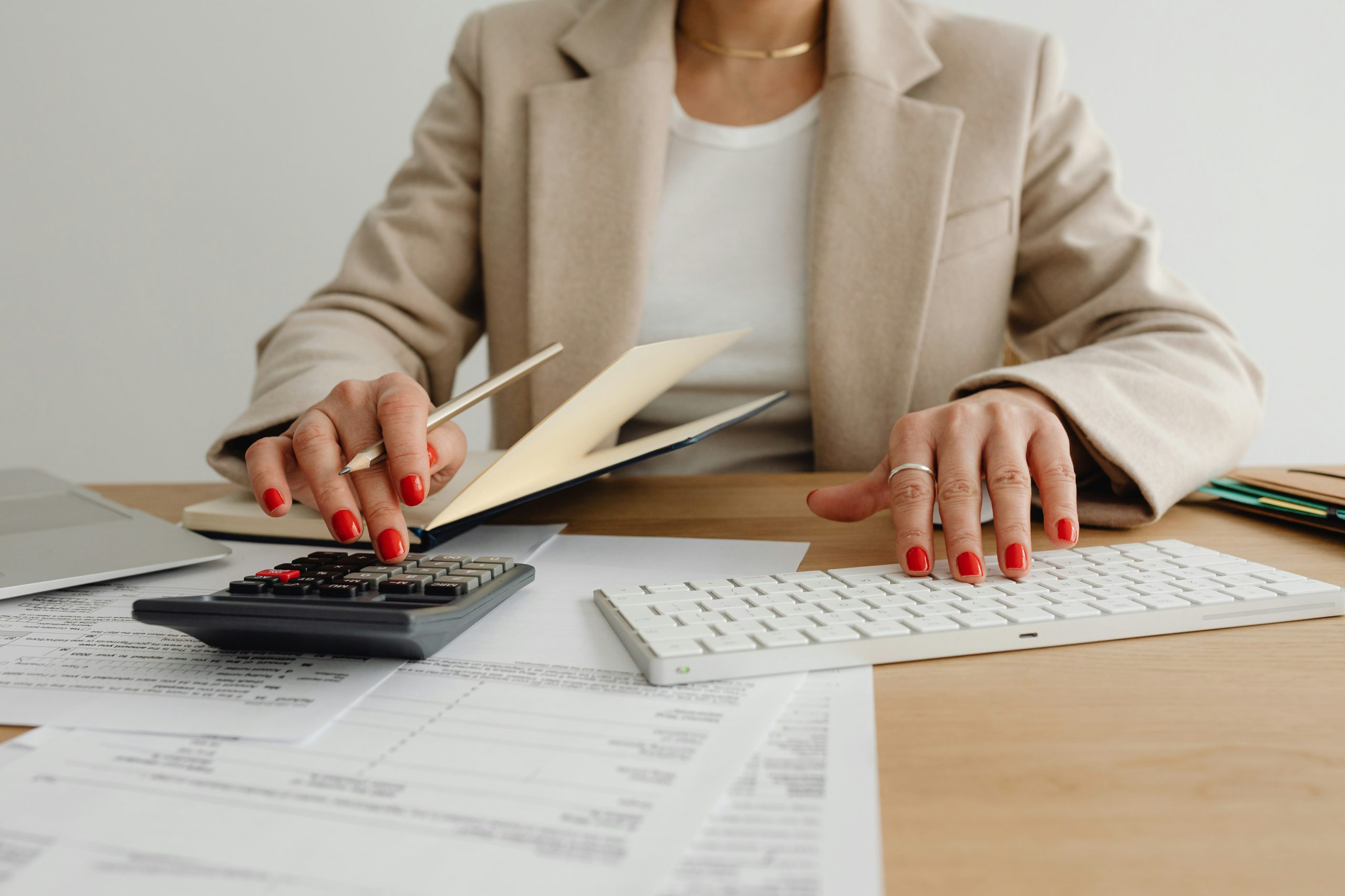 A person wearing a beige blazer and white shirt is sitting at a desk, using a calculator and a white computer keyboard. The desk has documents, a pen, and a folder. The person's nails are painted red.