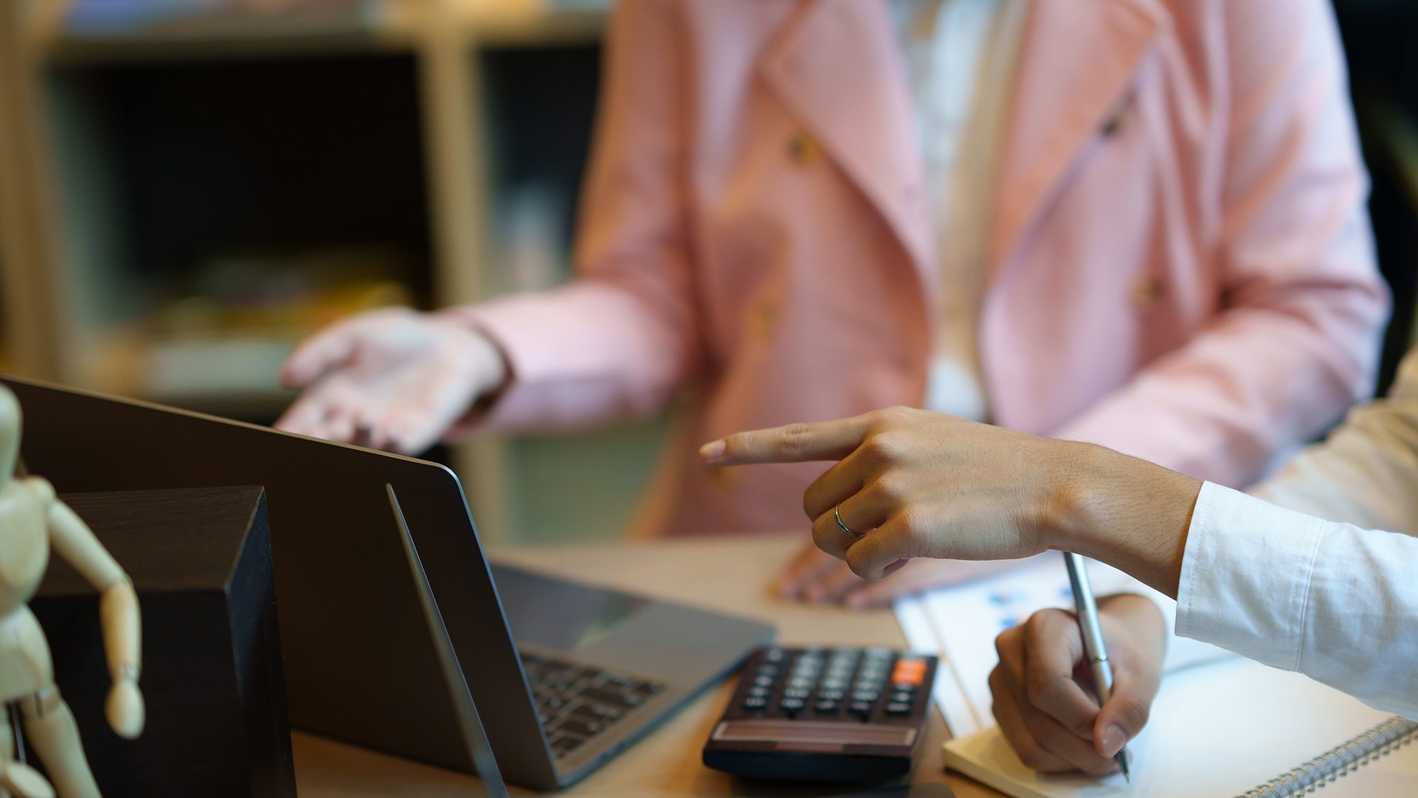 Two individuals are collaborating at a desk. One person is pointing at a laptop screen, while the other gestures with their hand. A notebook, calculator, and a small wooden mannequin are also on the desk. The focus is on their hands and the objects.