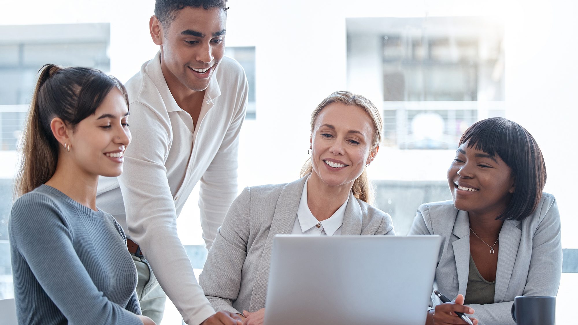 A group of four diverse coworkers smiling and collaborating in front of a laptop in a modern office. A young man stands while three women sit, all appearing engaged and focused on the discussion or project displayed on the laptop screen.