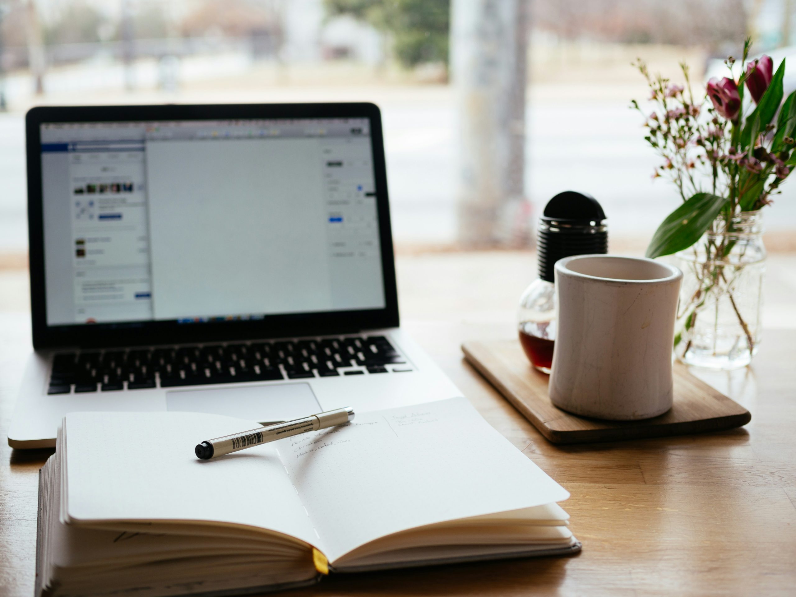 A wooden desk with an open laptop, a notebook with a pen, a white mug on a wooden coaster, and a small vase with flowers. The laptop screen shows a blurred document or web page. The background is blurred, showing an outdoor scene through a window.