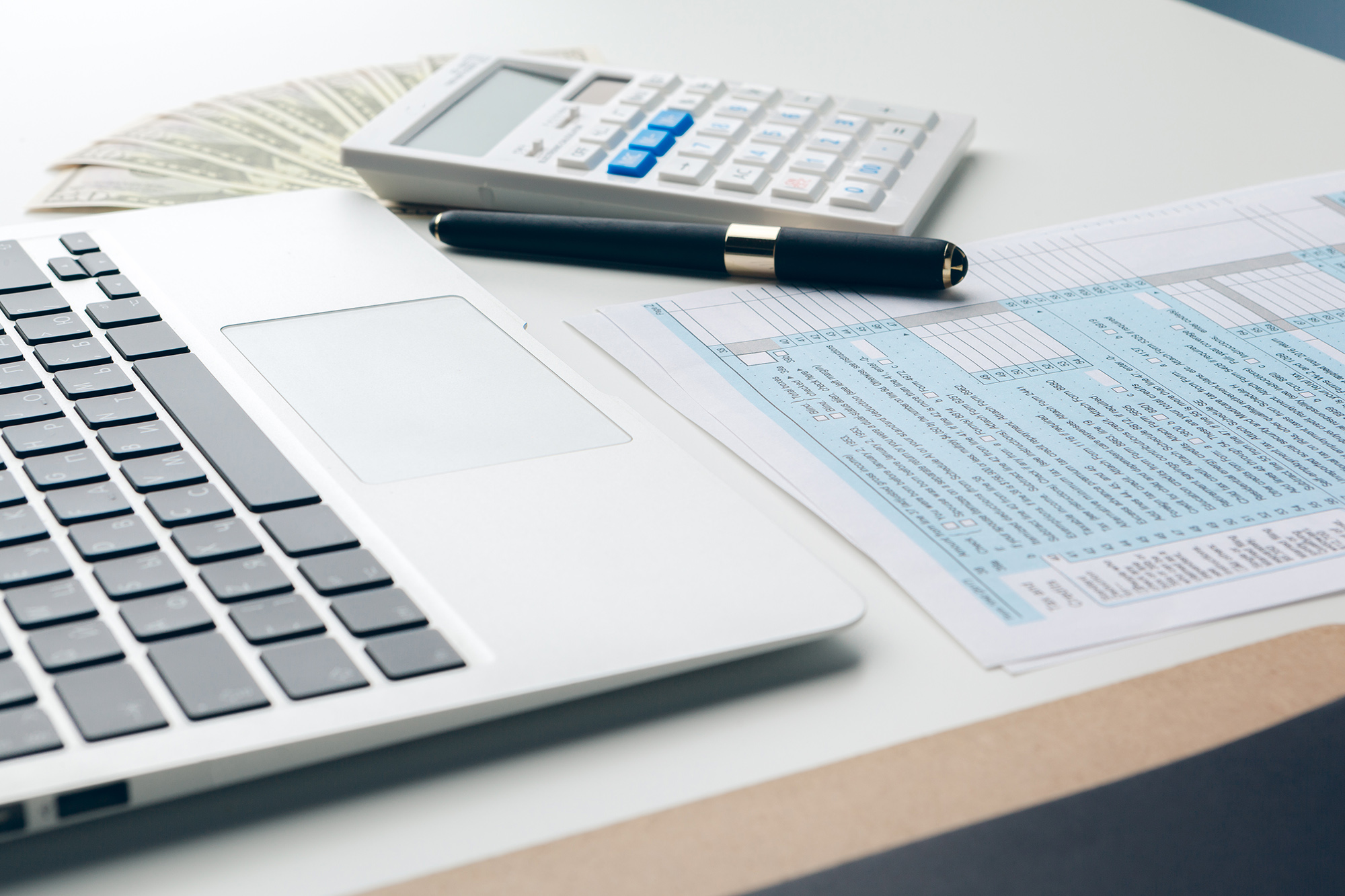 A close-up of a desk with a laptop, a black pen, a calculator, a stack of cash, and a tax form. The laptop's touchpad and part of the keyboard are visible. The pen is placed on top of the tax form, and the money is partially fanned out beside the calculator.