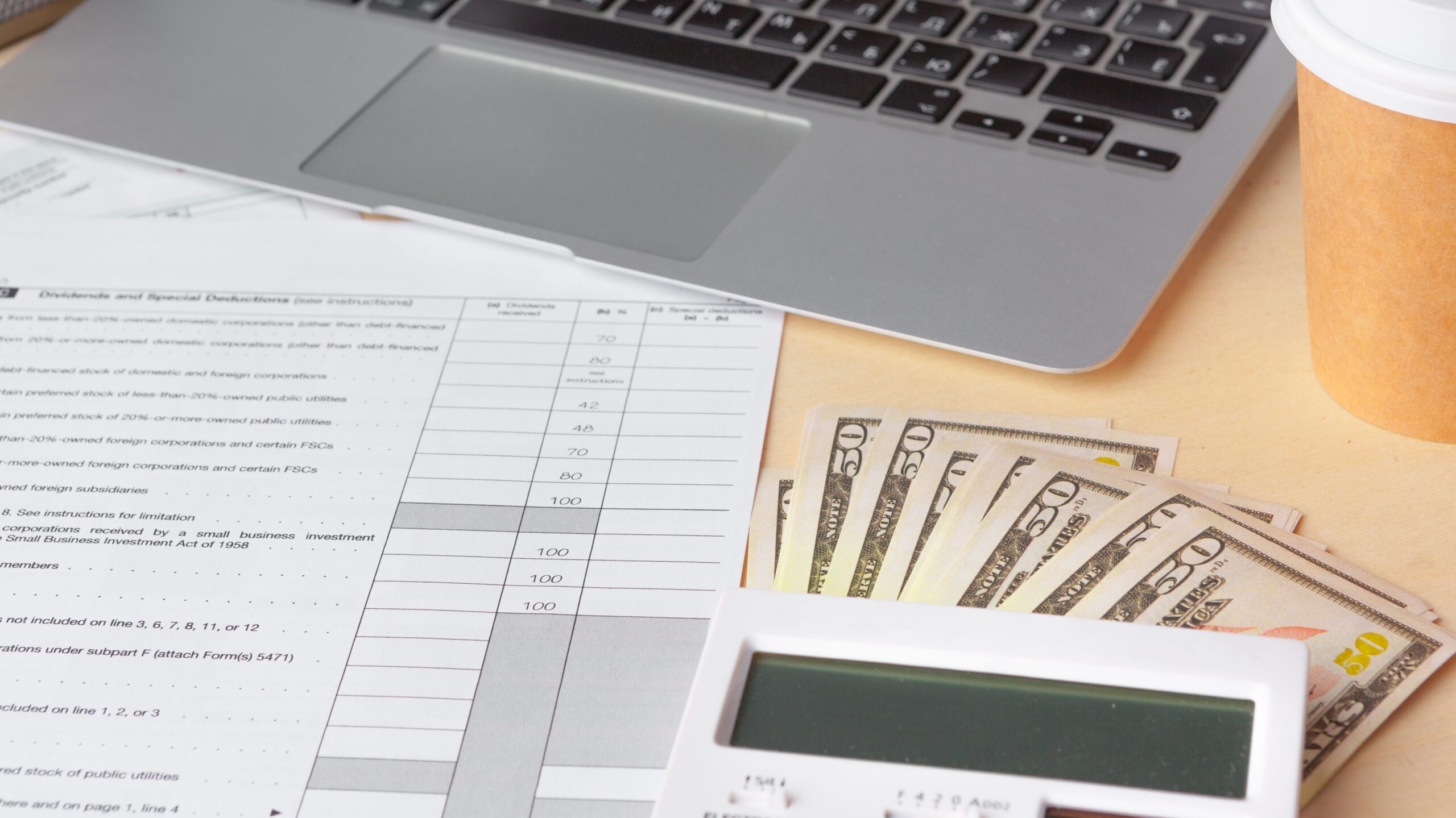 A laptop sits on a desk next to a filled-out financial form. Nearby, a few U.S. dollar bills are partially tucked under the form, and a white calculator is positioned in the foreground. A takeaway coffee cup sits on the right side of the desk.