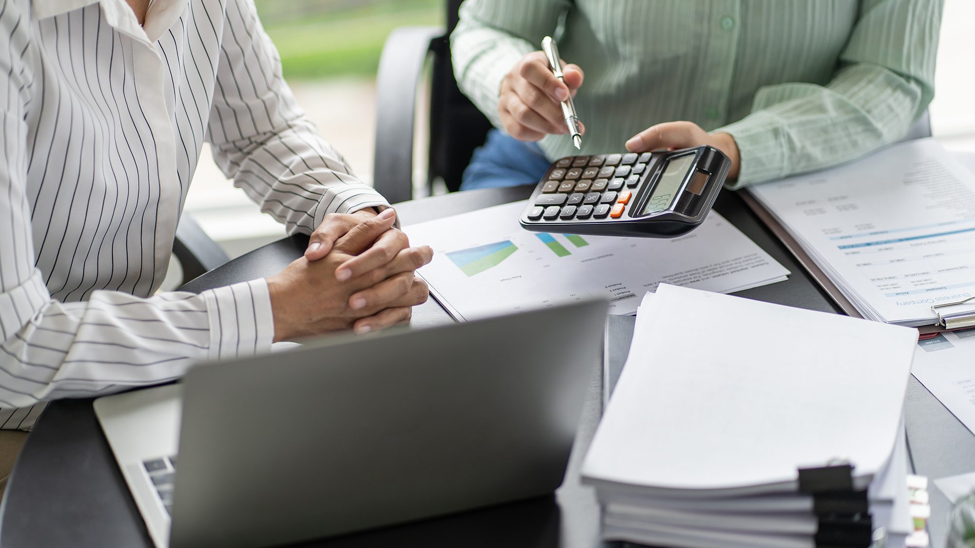 Two people collaborating at a desk cluttered with papers, documents, and a laptop. One person holds a calculator and points at it with a pen, while the other has hands folded. Charts and graphs are visible on some documents.