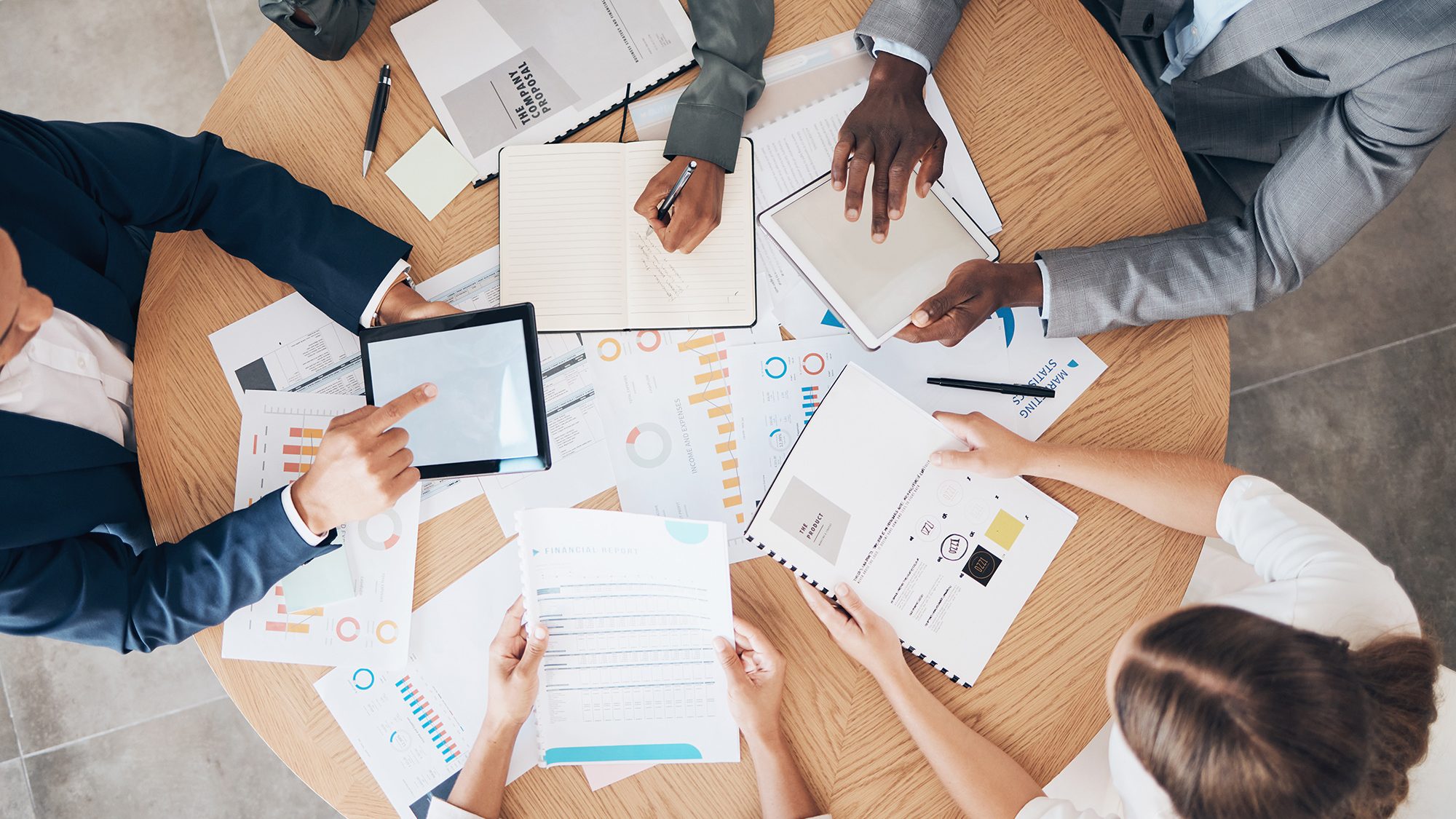 A top-down view of four individuals around a round wooden table engaged in a meeting or collaborative work session. Various documents, charts, graphs, and a tablet are spread across the table. The hands of the participants are seen gesturing and taking notes.