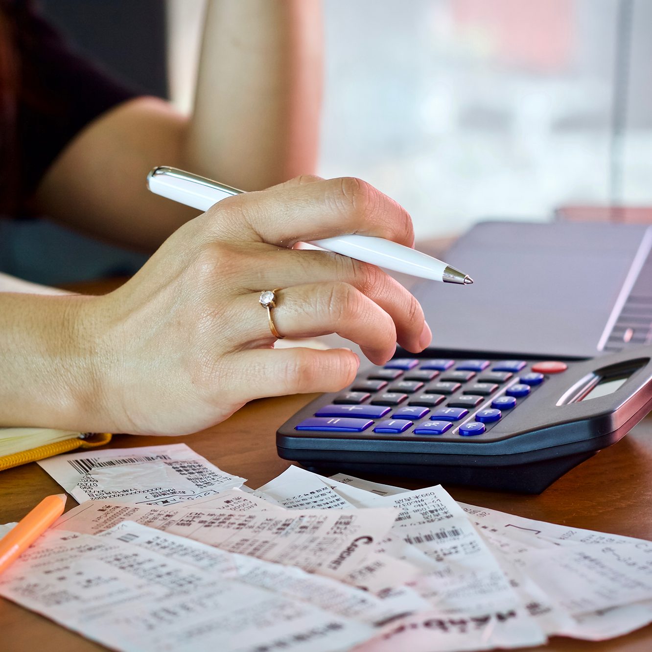 Close-up of a person's hand holding a pen, using a calculator. The person is wearing a ring on their ring finger. In the foreground, there are several receipts scattered on the wooden desk. A notebook and a yellow pen are also partially visible.