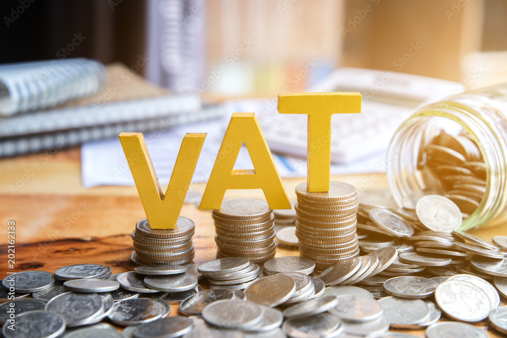 Stacks of coins support large, yellow letters spelling "VAT" on a table. A jar lies on its side, spilling more coins onto the surface. In the background, there are notebooks and papers, indicating a financial or business setting.