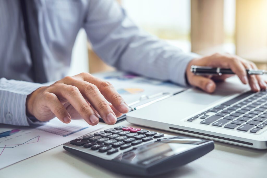 A person wearing a light blue shirt is using a calculator with their right hand and a laptop with their left hand. The person is holding a pen. The desk is cluttered with documents and charts. The scene appears to be an office or workspace setting.