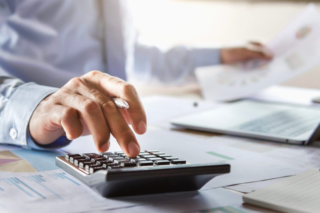 A person in a business shirt and tie uses one hand to press buttons on a calculator, while holding a document in the other hand. The surface is cluttered with papers, charts, and a laptop, suggesting a busy work environment.