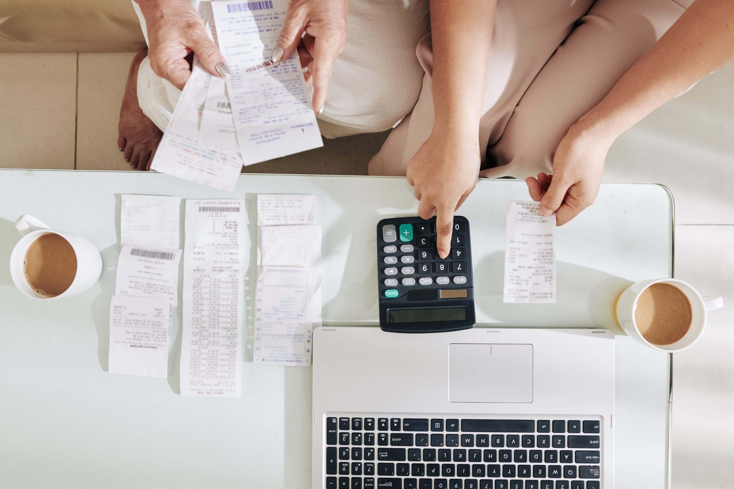 Two people are organizing receipts on a white table, with a laptop, a calculator, and two mugs of coffee. One person points to a receipt, and the other operates the calculator, indicating they are working on finances or budgeting.