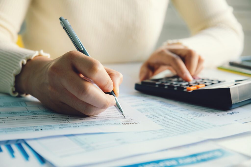 A person wearing a light-colored sweater is filling out forms with a pen in one hand and using a calculator with the other hand. Various documents and papers are spread out on the table in front of them.