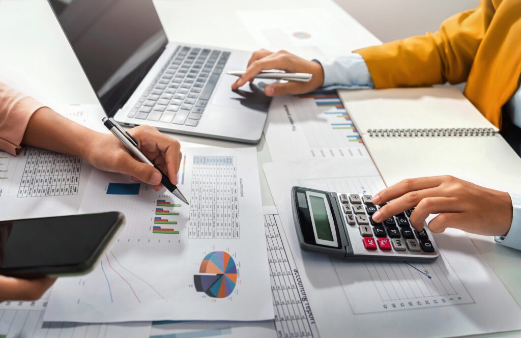 Two individuals working at a desk with a laptop, calculator, and various financial documents spread out. One person is holding a pen and checking charts, the other is using a calculator. A notepad is also visible on the table.