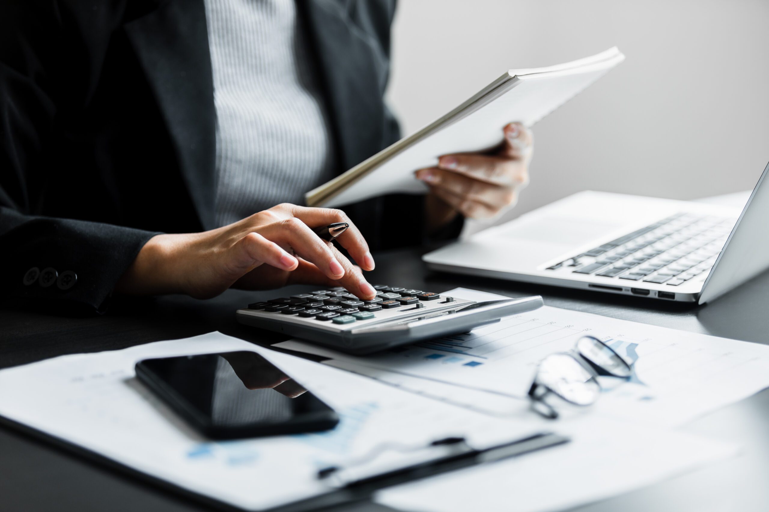 A person is working at a desk, using a calculator while holding a document. A laptop, smartphone, eyeglasses, and printed graphs are also on the desk. The individual is dressed in business attire, and the scene conveys a professional office setting.