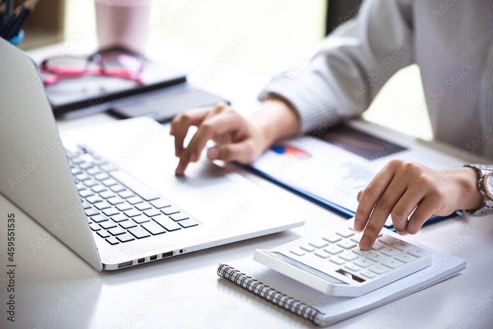 A person using a silver laptop with one hand while their other hand operates a white calculator. A notepad and a blue pen are on the desk, and a pair of glasses rest nearby. The scene suggests a work or study environment focused on calculations.