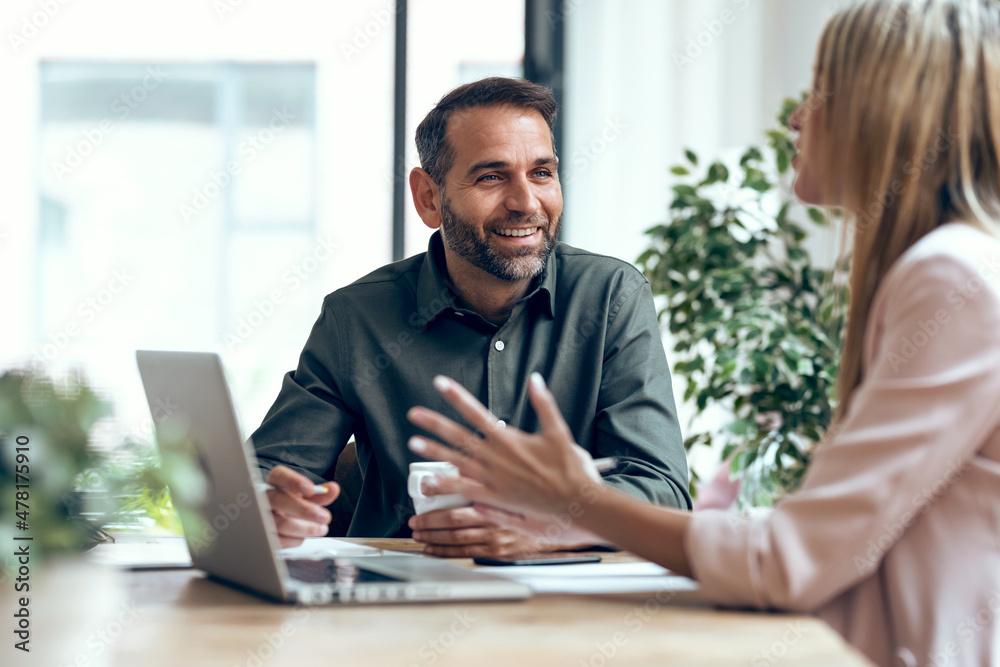 A man and woman sit at a desk with a laptop and papers. The man, wearing a green shirt, holds a pen and smiles, while the woman, with long blonde hair, gestures with her hands. They are engaged in a conversation in a bright, plant-filled office space.