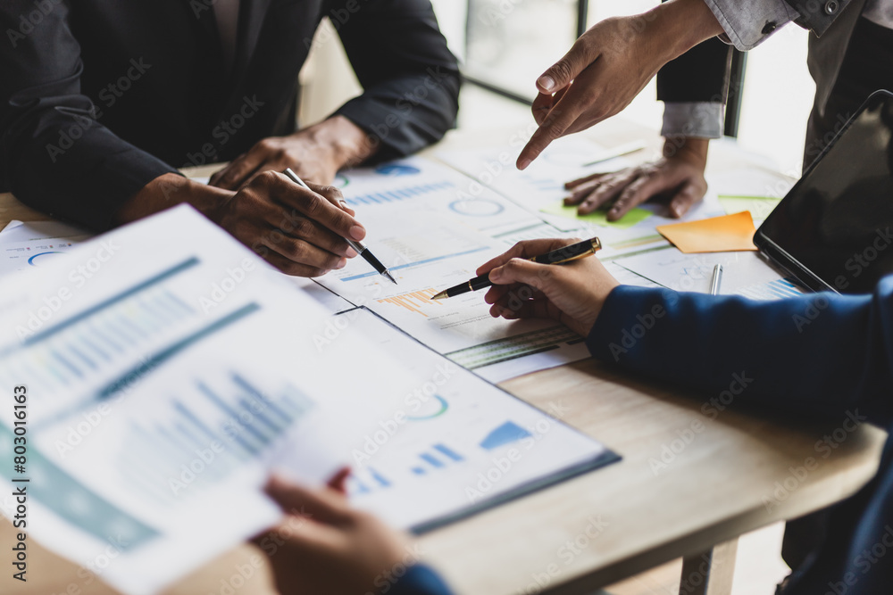 A close-up of a group of business professionals collaborating on a project. They are reviewing documents, charts, and graphs spread across a table. Hands are pointing and gesturing, indicating an active discussion.