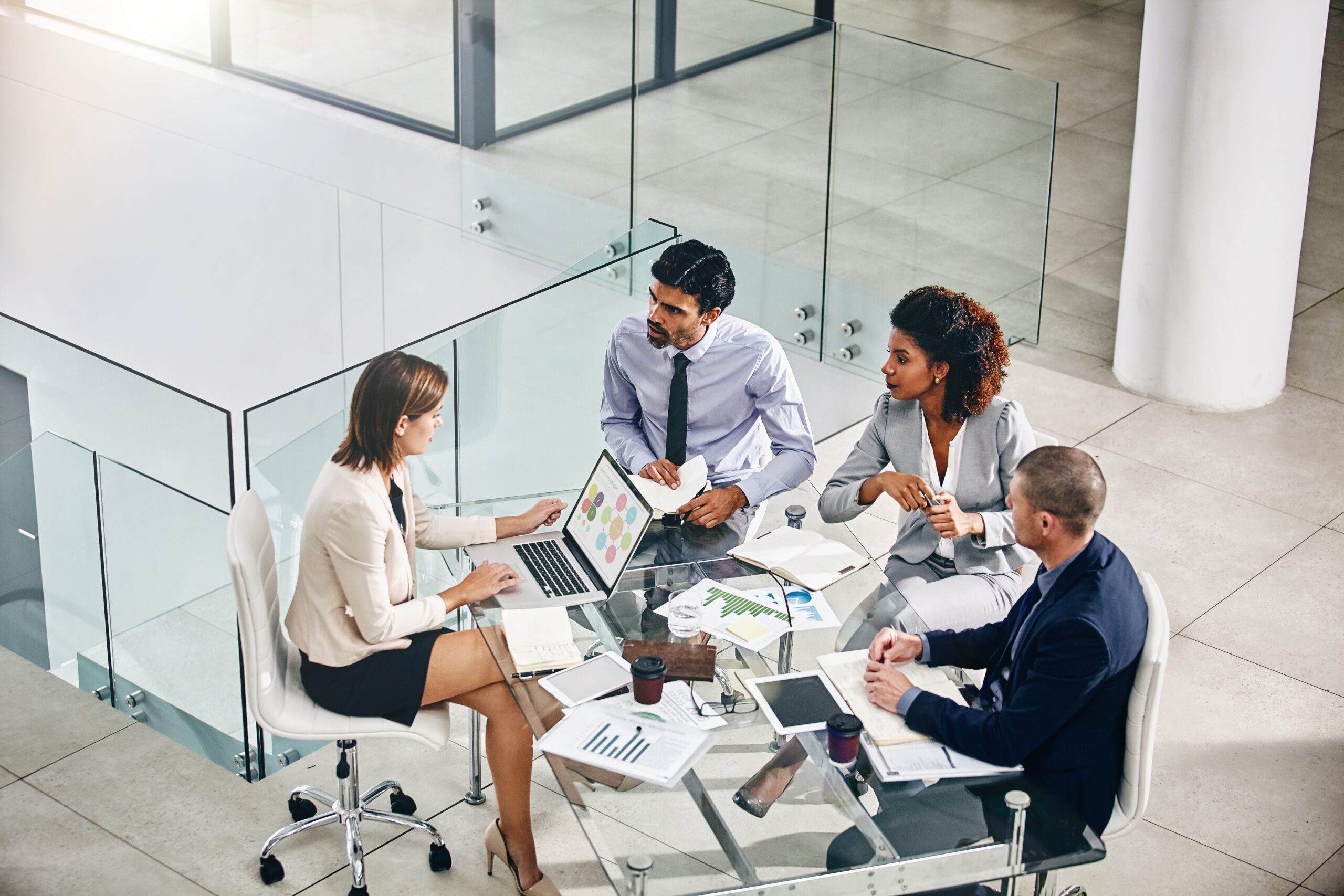 Four professionals are gathered around a round glass table in a modern office. They are engaged in discussion, with one person typing on a laptop while another shows a document with colorful charts. Papers, tablets, and coffee cups are scattered on the table.