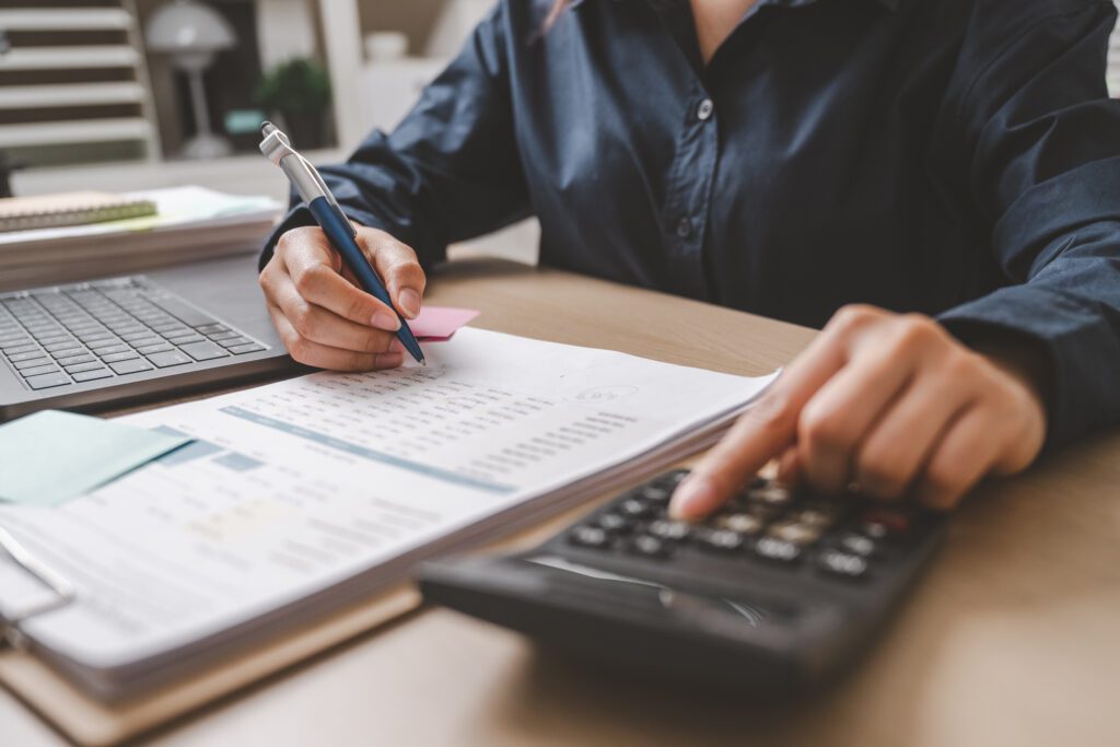 Person using a calculator and writing on a financial document at a desk, with a laptop and sticky notes nearby.