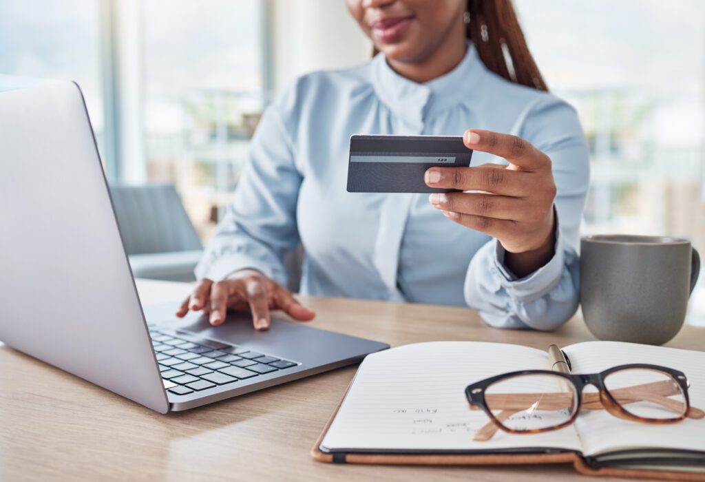 A woman sits at a desk using a laptop, holding a credit card in one hand. A notebook with glasses and a cup are also on the desk, suggesting she is shopping or banking online.