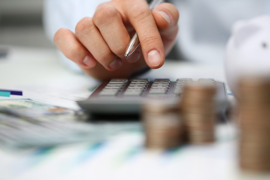 A close-up of a hand pressing buttons on a calculator with a pen, next to stacked coins and some blurred banknotes, suggesting financial calculations or budgeting.