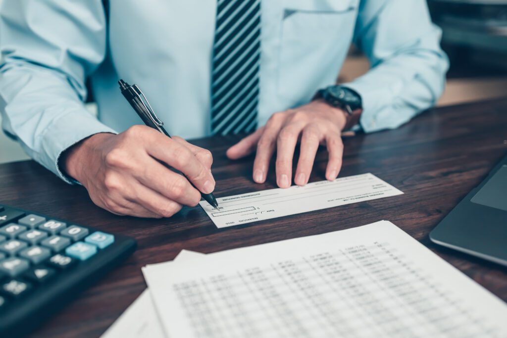 A person in a blue shirt and striped tie sits at a desk, writing on a check. A calculator, financial documents, and part of a laptop are also visible on the desk.