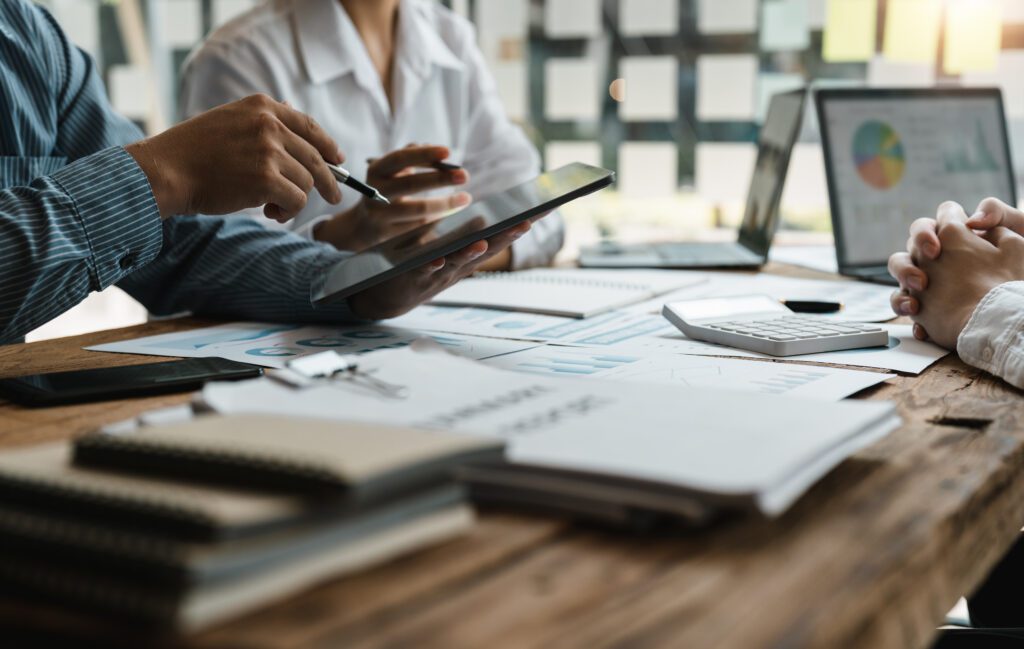 Three people sit at a table covered with documents, a tablet, laptop, calculator, and charts. One person points at the tablet, while others listen, suggesting a collaborative business meeting or discussion.