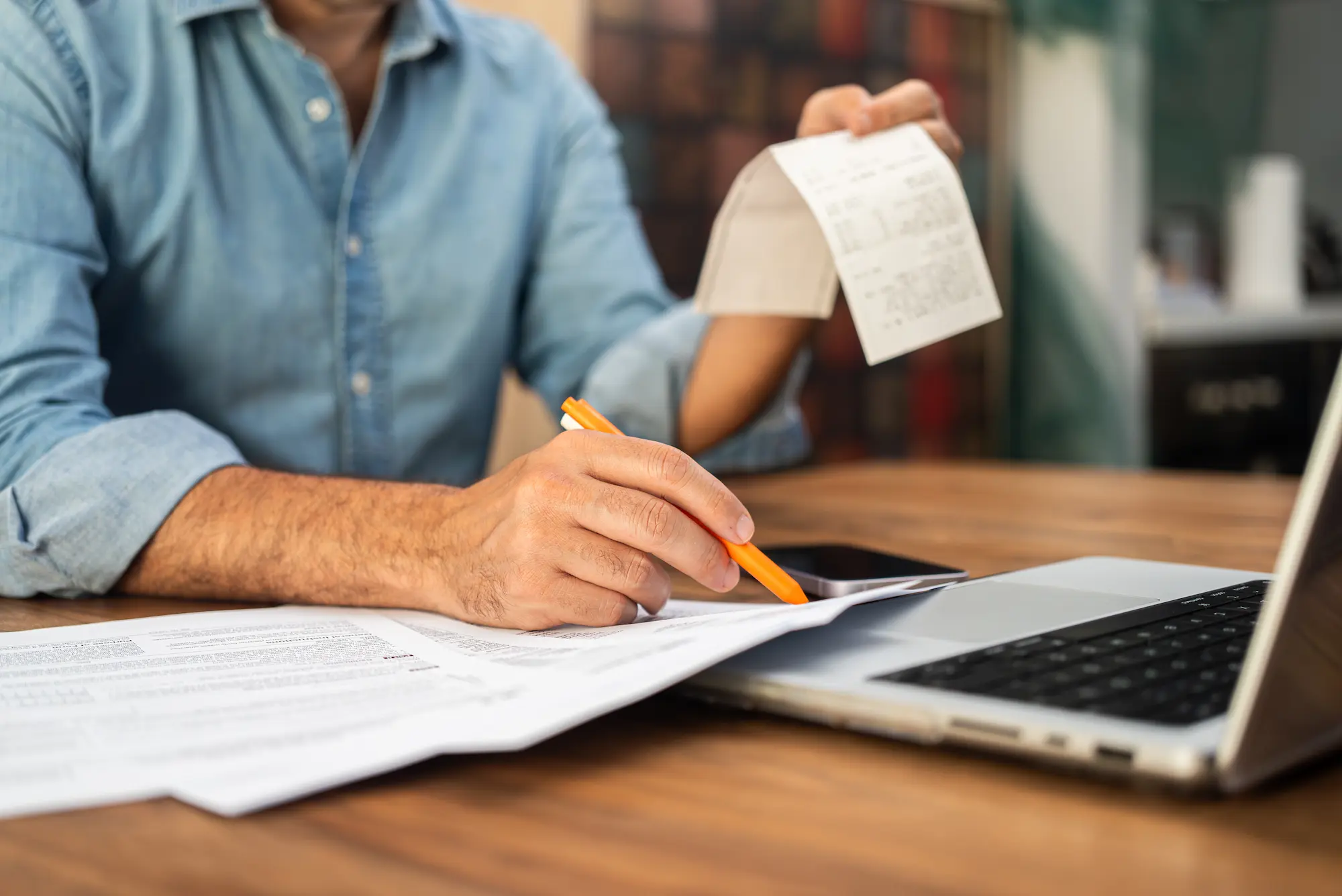 A person in a denim shirt reviews bills and receipts at a wooden table, using a pencil to write on papers beside a laptop.