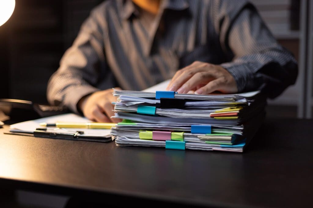 A person in a striped shirt reviews a large stack of documents organized with colorful binder clips and sticky notes, sitting at a desk under a lamp.