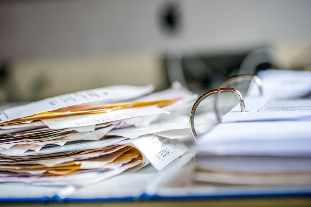 A close-up of a messy stack of papers and receipts next to an open ring binder on a desk, with a blurred background.